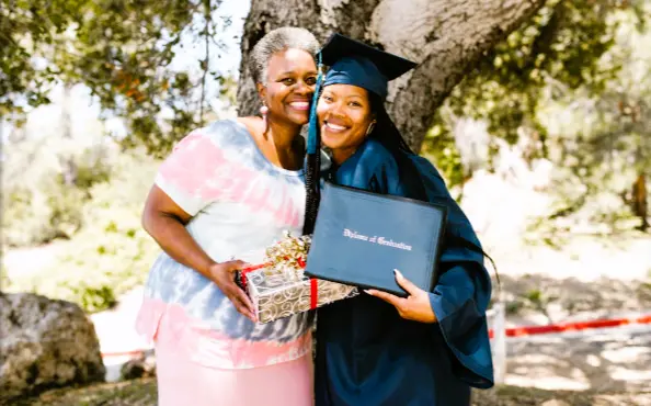 A young graduate holding her diploma and wearing her graduation gown and cap next to a friend